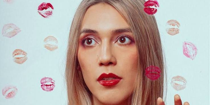 Close-up of a blonde woman with red lipstick against a pale blue wall, surrounded by lipstick kiss prints, looking surprised.
