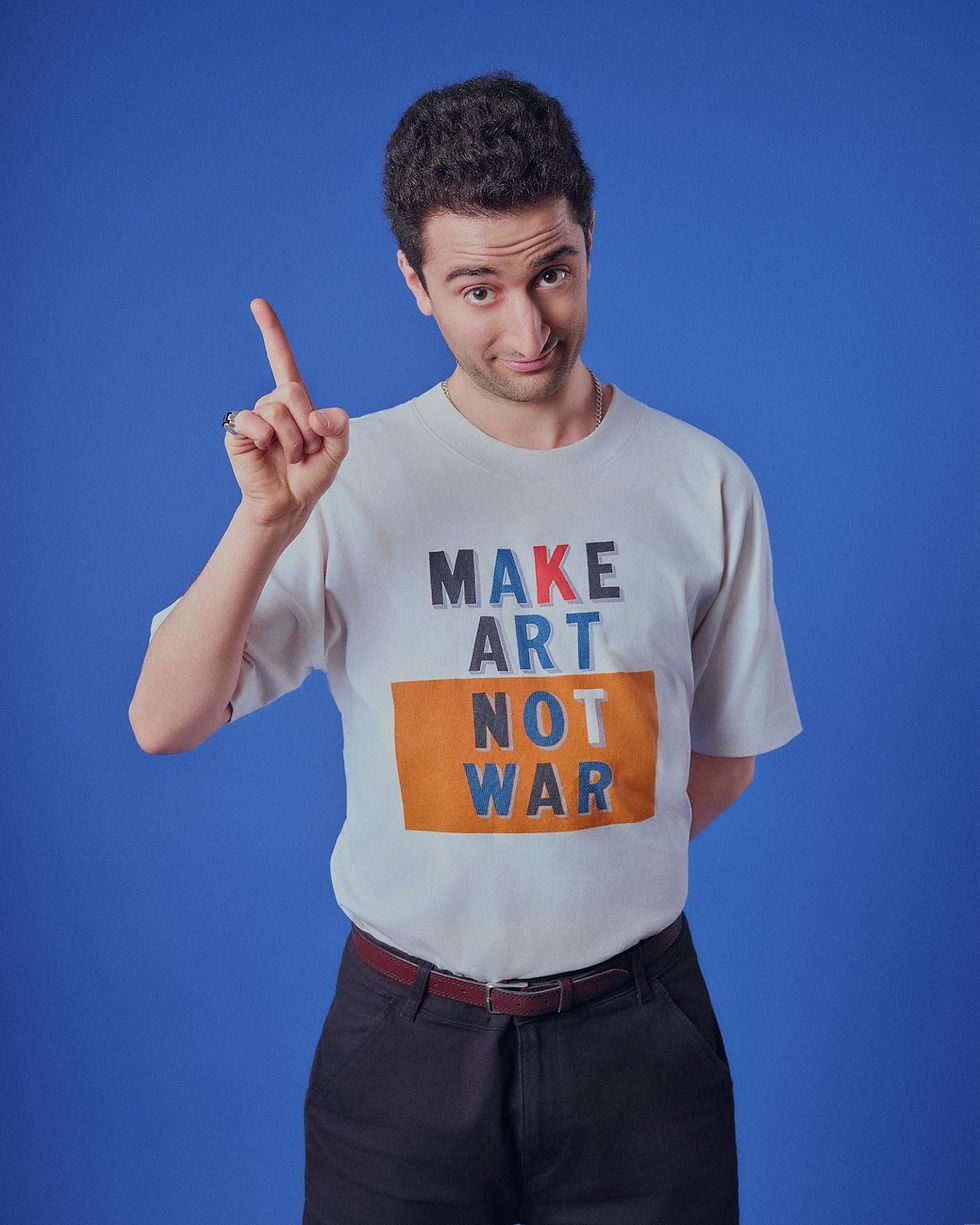 Man in white MAKE ART NOT WAR T-shirt points up against a blue studio backdrop, with a confident, playful expression.