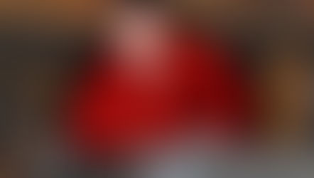 Man in ornate red outfit crouches on an old ship next to a cannon, amid wooden beams and barrels, exuding focus and determination.