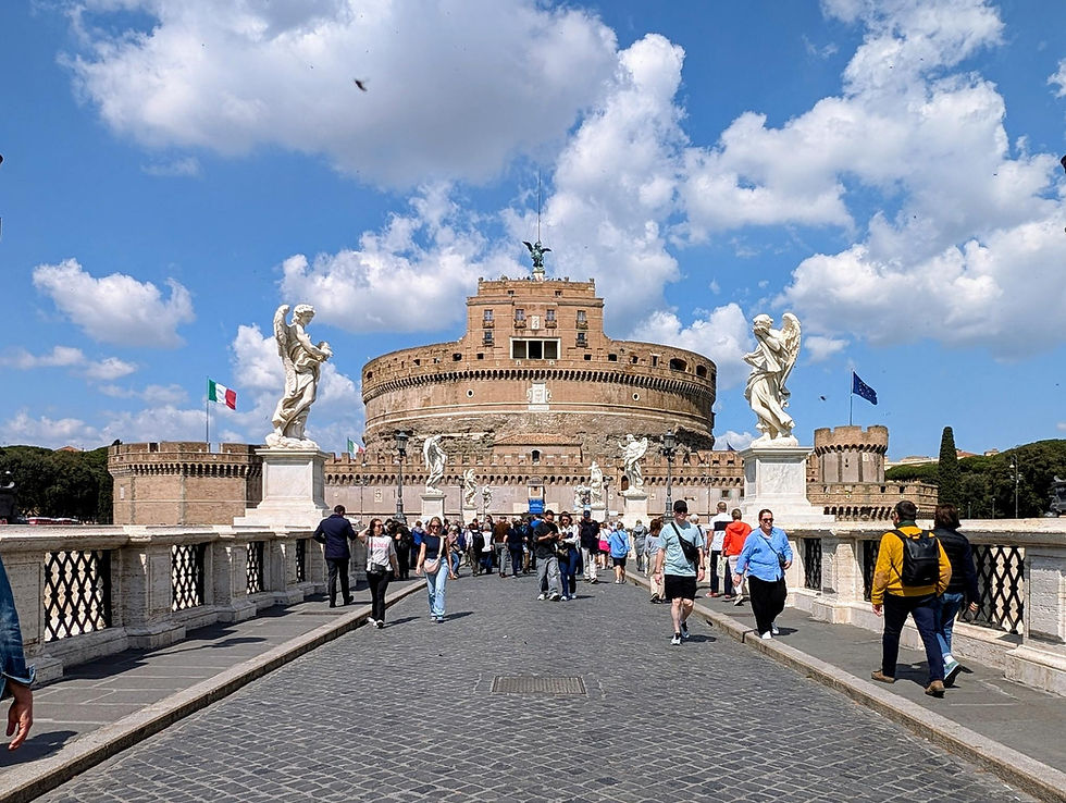 People walk on a stone bridge toward Castel Sant'Angelo in Rome. Statues and flags are visible under a bright blue sky with clouds.