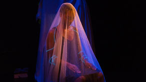 Veiled woman seated on a dark stage, lit in blue and purple, with glowing blue bracelets and a quiet, solemn mood.
