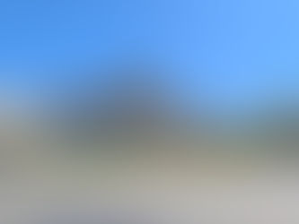 Stone arch monument with ornate sculptures in a sunny plaza. Surrounding it are colourful flowers, greenery, and a clear blue sky.