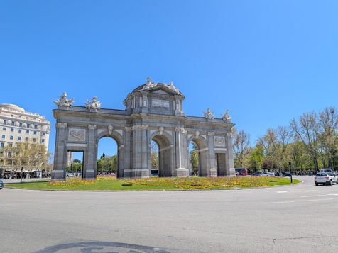 Stone arch monument with ornate sculptures in a sunny plaza. Surrounding it are colourful flowers, greenery, and a clear blue sky.