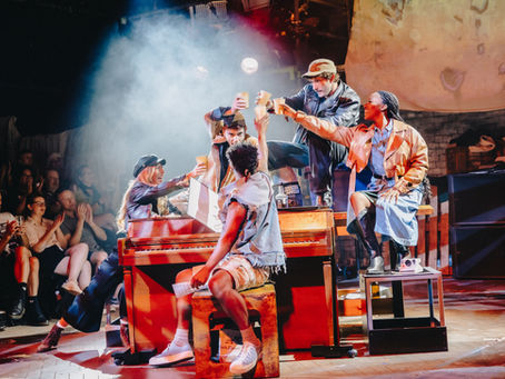 Actors on a smoky stage raise paper cups around a piano, while audience members watch and applaud in warm red light.