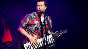 Musician in a colourful shirt plays a keytar on stage, singing into a microphone. A dimly lit background with a faint clock is visible.