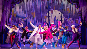Performers in colorful costumes dance energetically on a vibrant stage with a whimsical, snowy backdrop. Mood is joyful and festive.