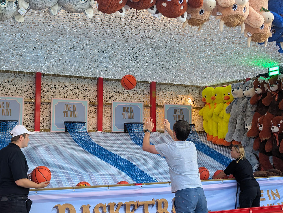 Carnival basketball game with people shooting orange balls toward nets beside plush duck and bear prizes; One in to win signs.
