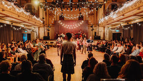 Audience watches a lively folk concert in a warm-lit ballroom, with musicians playing on stage and strings of lights overhead.
