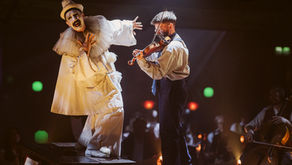 A clown in white costume, face expressive, gestures dramatically near a violinist playing on stage. Dimly lit background with warm hues.