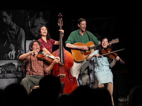 Four smiling musicians perform on a dark stage with fiddle, guitar, and upright bass, backed by black-and-white photos.