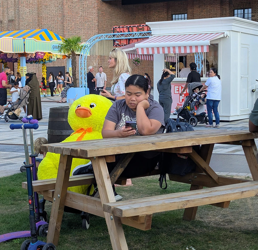 Crowded outdoor fair with a woman using her phone at a picnic table beside a giant yellow duck plush and ice cream kiosk.