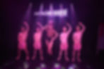 Performers on stage with pink hats, one in drag attire, under purple lights. Sign reads "Boys in the Buff." Playful mood.
