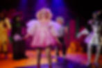 Panto fairy on stage in a pink dress with other characters from Oz/Wicked in the background