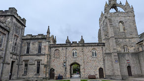 Stone castle courtyard with Gothic tower and archway, green lawns, cloudy sky, and a few people near the centre entrance