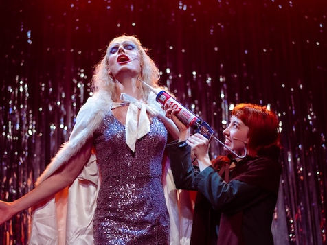 Drag artist in sequin dress and white faux fur cape standing, with a woman applying filler to their neck