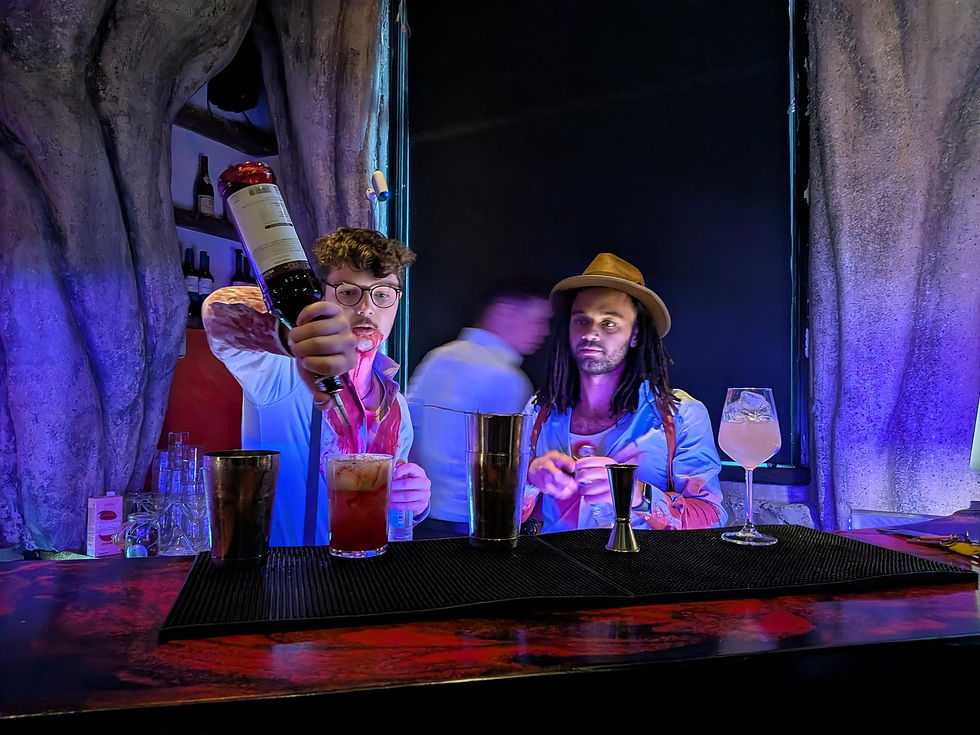 Two bartenders mix cocktails behind a dark stone bar, one pouring wine, under blue-purple lighting with glasses and shakers