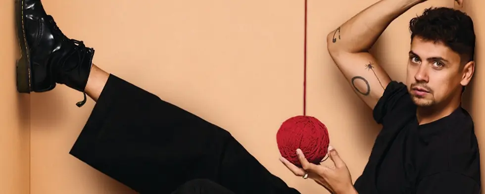 Man in black poses against a beige wall, one leg up, holding a red yarn ball; moody fashion portrait.