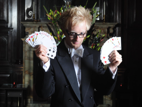A man wearing a tux and a white bow tie holds a pack of cards in each hand