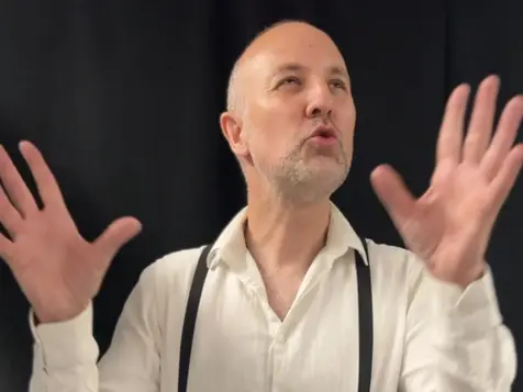 Bald man in a white shirt and braces gestures with raised hands against a dark background, looking upward in surprise.
