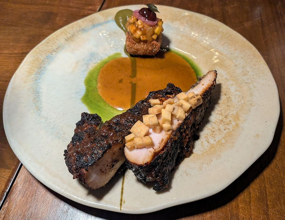 Grilled meat with diced mangoes on a plate, green sauce, and a small garnished cube. Brown wooden table surface.