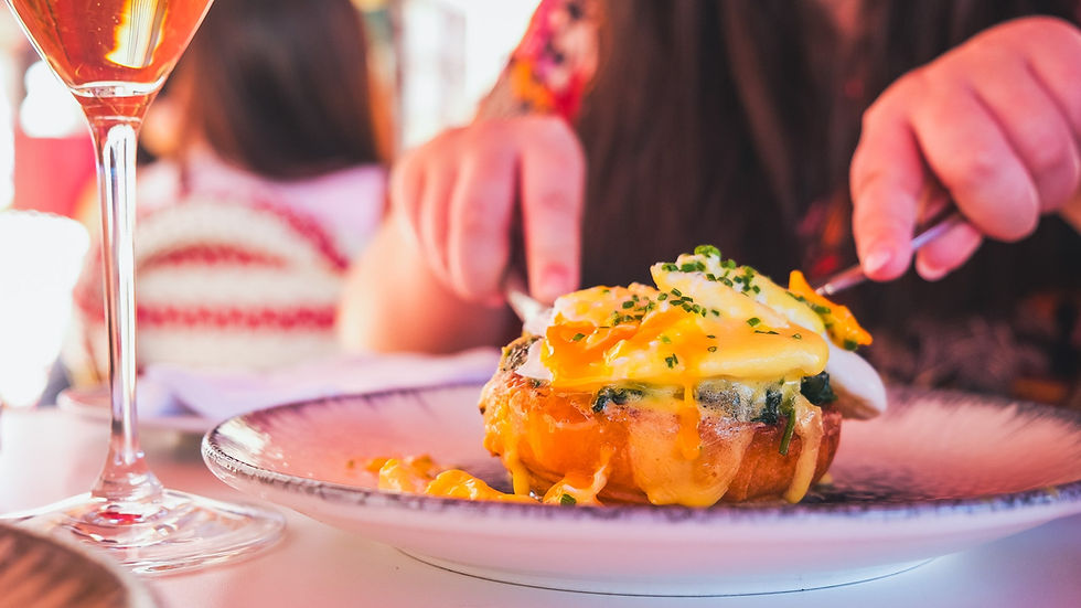 A person cuts into eggs Benedict with hollandaise sauce at a restaurant. A glass of orange drink is nearby. Relaxed dining atmosphere.