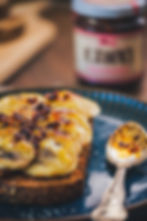 Toast with banana slices and crispy toppings on a blue plate, alongside a spoon. Blurry jar in background suggests a kitchen setting.