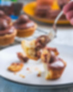 A fork holds a bite of a chocolate-topped muffin on a plate. Crumbs surround it. Background shows more muffins on a wire rack.