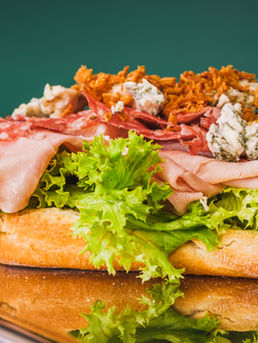 Large sandwich with meats, cheese, and lettuce on a bun against a green background. Text: Deli and Co., custom sandwich spot.