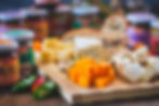 Assorted cheese cubes and wedges on a wooden board with green and red peppers. Jars with colorful labels in the blurred background.