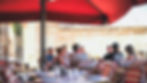 People sit at outdoor cafe under a red umbrella, engaging in conversation. Tables with drinks and striped chairs create a relaxed vibe.
