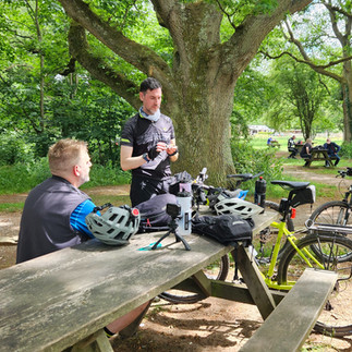 Cyclists in gear gather at a wooden picnic table in a green park, adjusting equipment. Bright helmets and bikes nearby; relaxed, outdoorsy vibe.