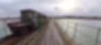 Old train on a narrow pier beside calm water, under cloudy skies. British flags decorate the train. Houses visible in the background.