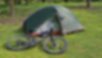 A green tent and a black bicycle with striped handles are on lush grass in a wooded area, suggesting a camping scene.