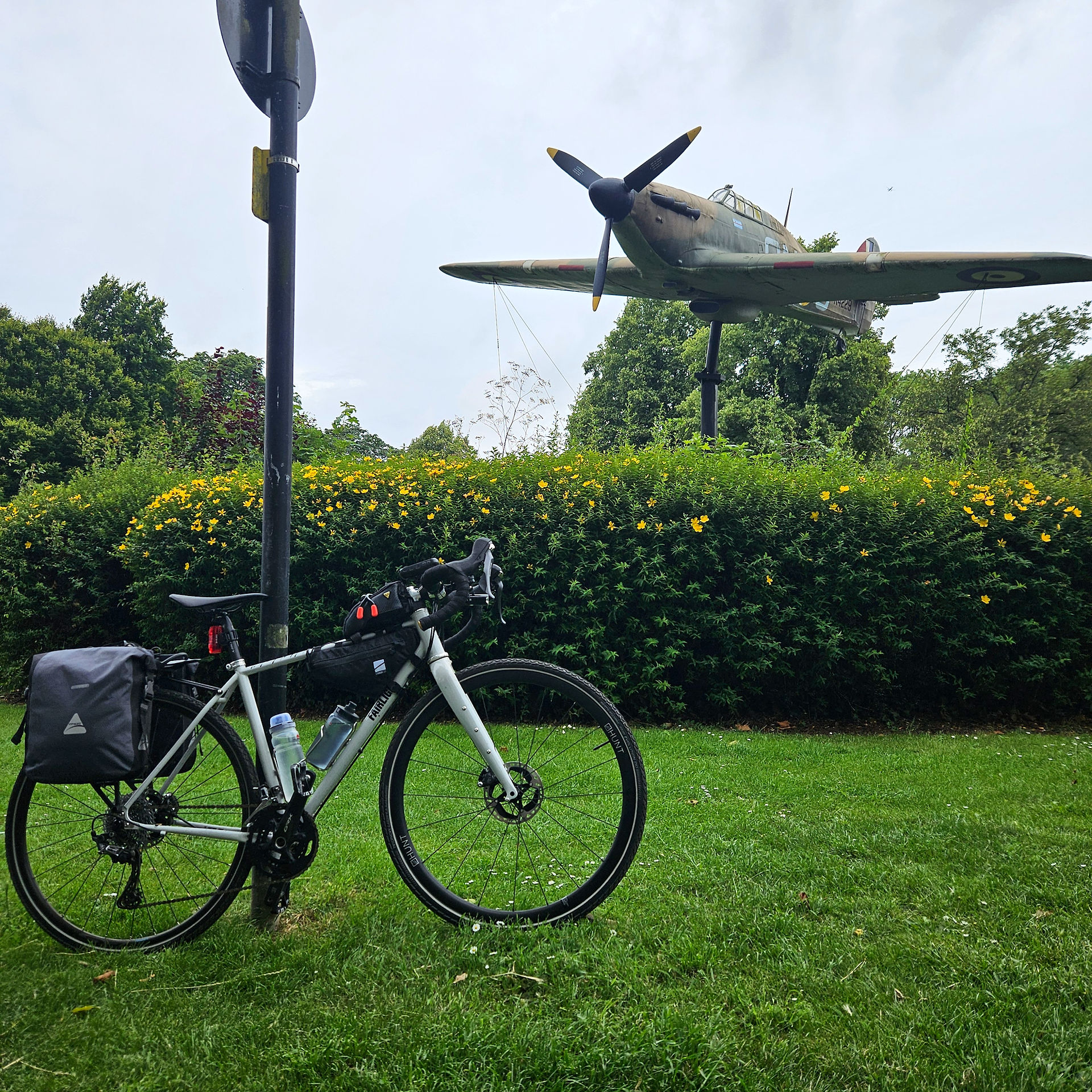 A bike with panniers propped against a post with hurricane world war 2 fighter jet in the background