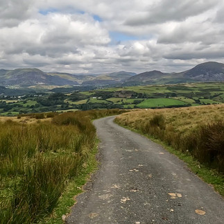Winding road through grassy hills under a cloudy sky, leading to distant green fields and mountains. Serenity and natural beauty abound.