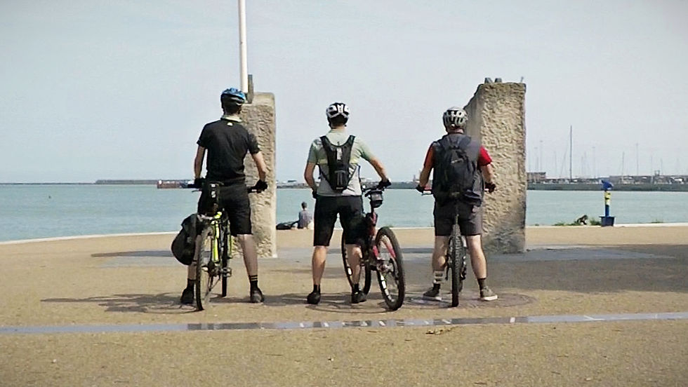 Three cyclists in helmets face the sea on a sunny day, standing beside their bikes on a paved path, with a marina in the background.