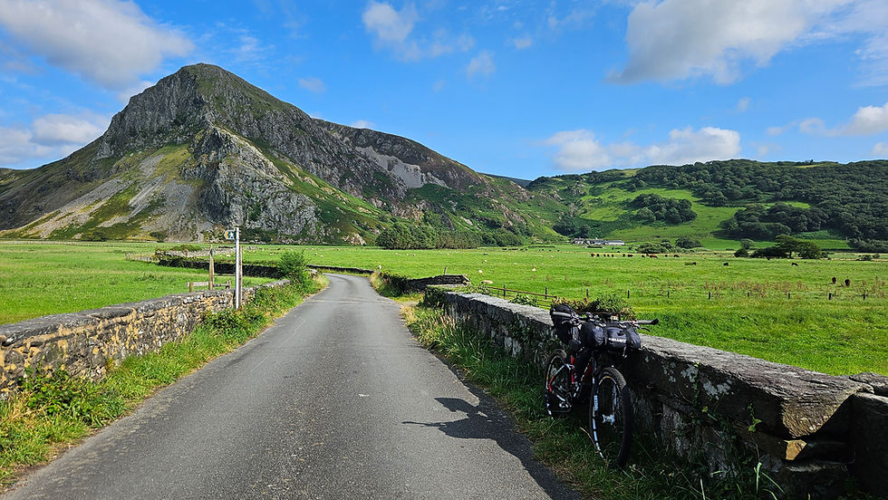 Mountain landscape with a road and bicycle leaning on a stone wall. Green fields and cattle under a blue sky with clouds in the background.