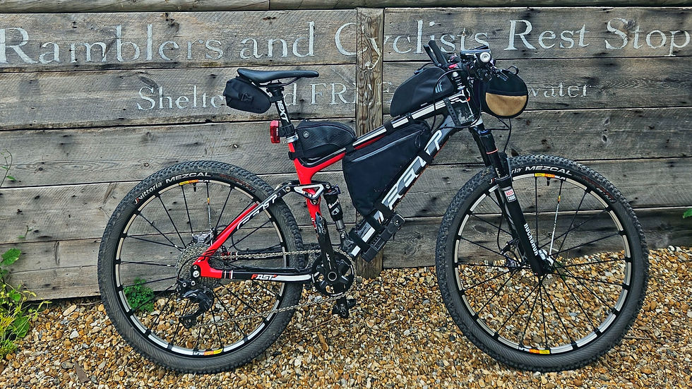 Red and black bike parked against a wooden fence with text "Ramblers and Cyclists Rest Stop." Gravel ground, lush green foliage.