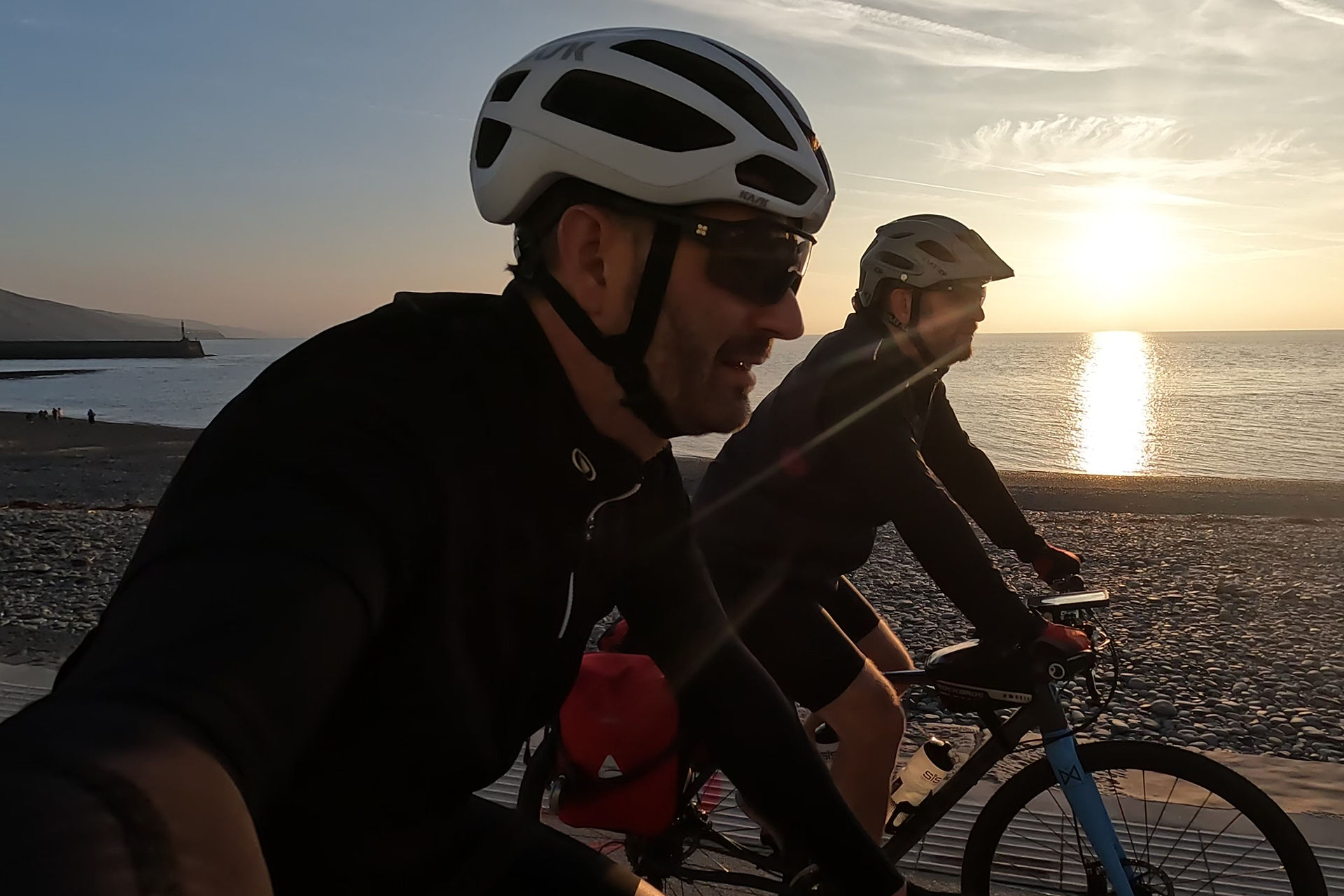 Two cyclists ride along a beachside path at sunset, wearing helmets and sunglasses. The sun reflects on the calm sea, creating a serene mood.