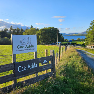 A sign post to a campsite with lush green fields adn trees in the background. Text says Cae Adda