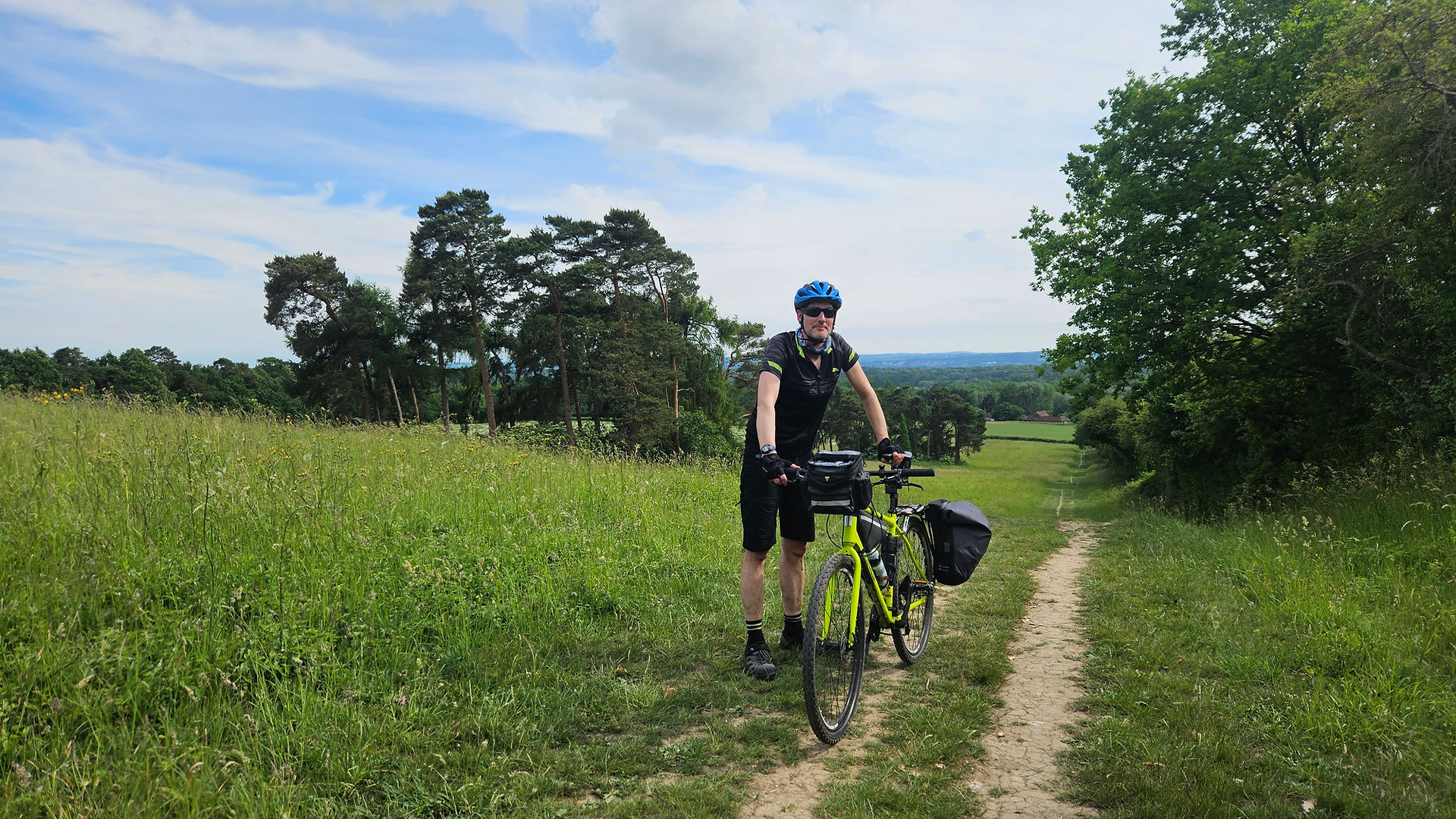 Cyclist in blue helmet stands with a yellow bike on a grassy path. Trees and blue sky in the background create a serene setting.