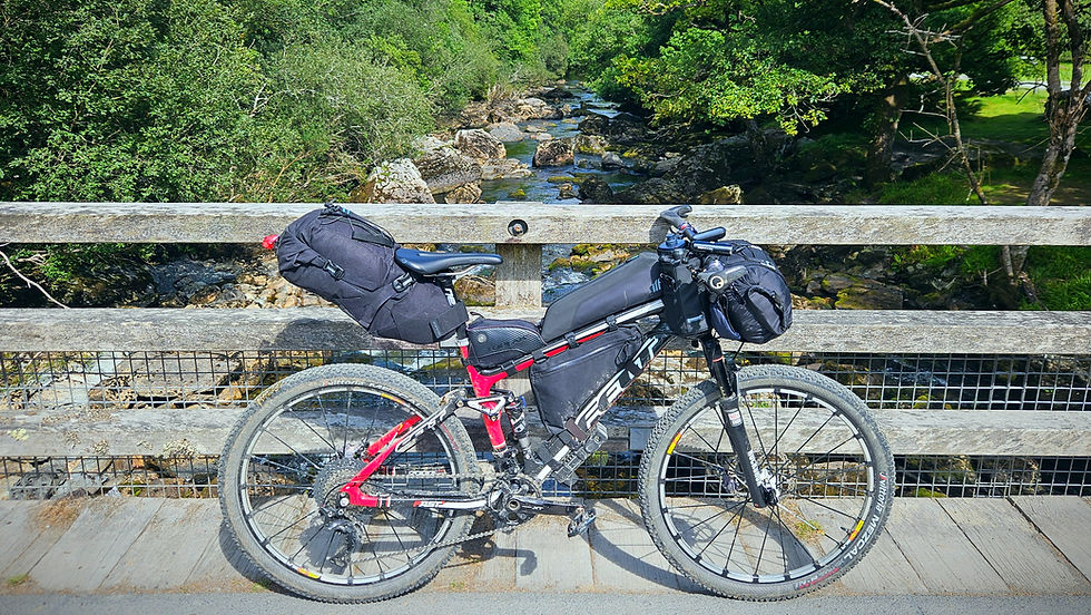 Bicycle with bags on a wooden bridge, overlooking a rocky stream. Lush green foliage surrounds, creating a serene outdoor setting.