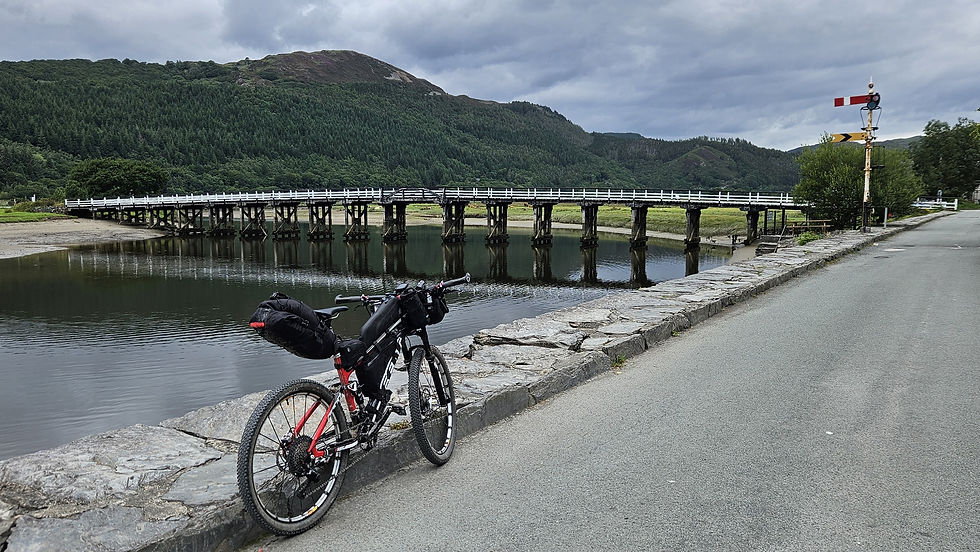 Bicycle parked by a stone pathway, with a wooden bridge spanning a reflective river. Lush green hills and cloudy sky in the background.