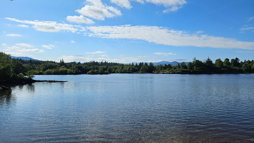 Calm lake with clear blue sky, scattered clouds, and distant tree-covered hills. Peaceful, natural landscape setting.