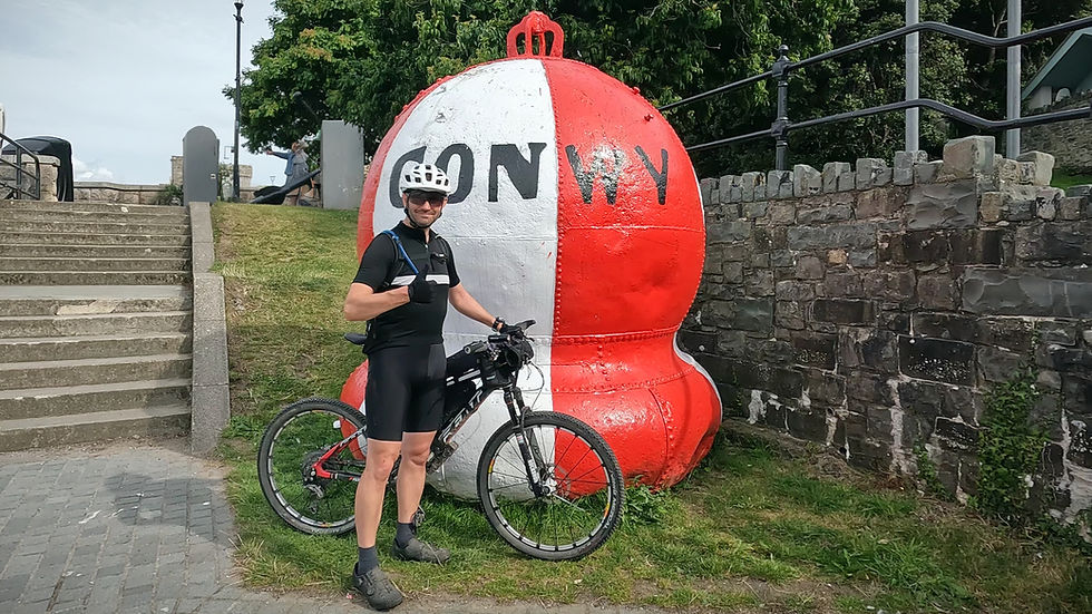 Cyclist poses with thumbs up beside bike and large red-and-white buoy with "CONWY" on it, near stone wall and grassy area. Sunny day.