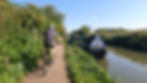 Cyclist rides along a canal path next to a blue narrowboat. Greenery lines the path under a clear blue sky, evoking a peaceful atmosphere.