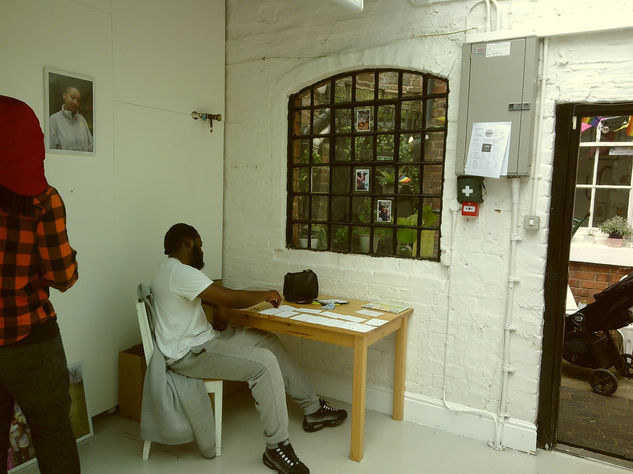 A man sits at a table near the entrance window, sticking blue tack to small photographs. The back of another man can be seen as he hangs portraits.