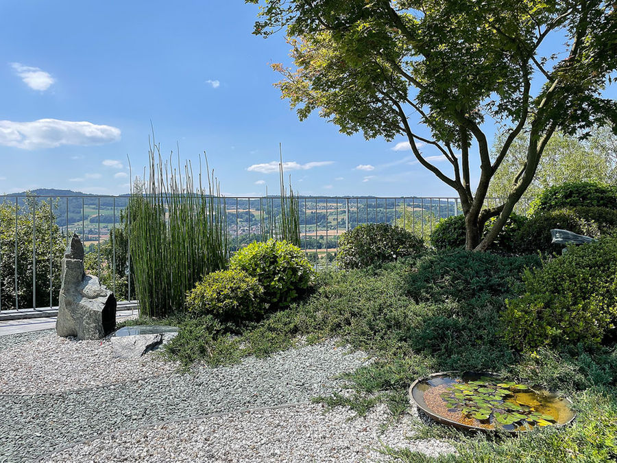 Japanese-style garden with sculptural stones, horsetail reeds, Japanese maple, evergreen groundcover and a water bowl, creating a calm Zen-inspired landscape with panoramic hillside views.
Japanisch gestalteter Garten mit skulpturalen Steinen, Schachtelhalmen, japanischem Ahorn, Immergrün und Wasserschale – eine ruhige, zenhafte Landschaft mit weitem Hügelblick.