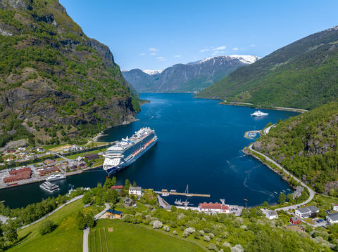 Aerial view of a fjord with a large cruise ship docked near a small village, surrounded by towering green mountains and snow-capped peaks.
