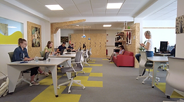 Menschen sitzen an Schreibtischen in einem Büro mit gelbem und grauem Bodenbelag.